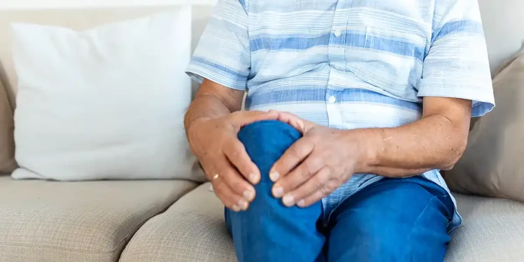 A close-up of an older adult's hands clasping their knee, suggestive of joint or bone discomfort, while seated on a couch, and he needs Hormone Therapy and Osteoporosis.