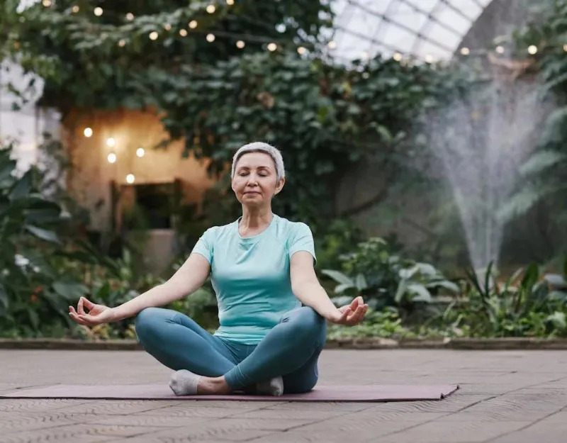 Vaginal Health: An older woman practicing yoga with a serene expression in a lush greenhouse setting.